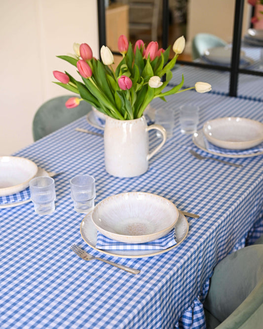 Gros plan sur une nappe à carreaux bleu et blanc avec volants, en coton 100 % fait main à Biarritz, dressée avec une vaisselle artisanale et des serviettes assorties. Au centre, un bouquet de tulipes rouges et blanches dans un pichet en céramique blanche.