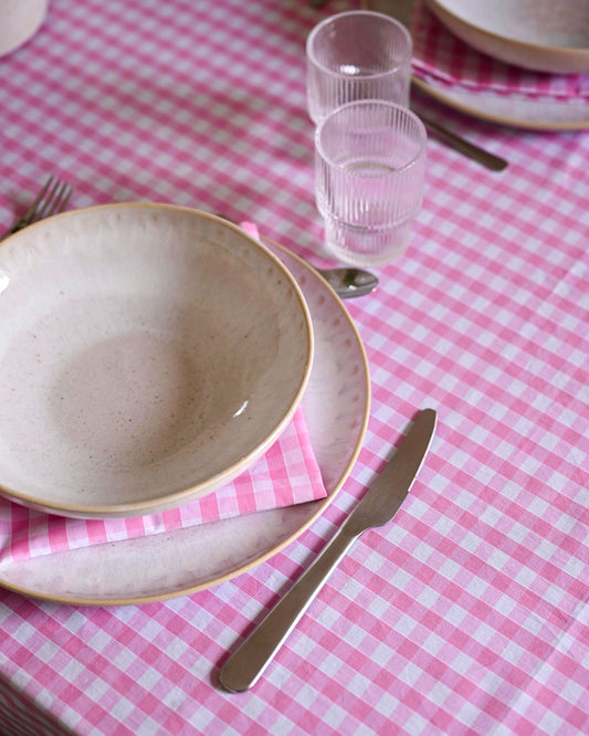 Gros plan sur une nappe à carreaux rose et blanc avec volants, en coton 100 % fait main à Biarritz, dressée avec une vaisselle artisanale et des serviettes assorties. Au centre, un bouquet de tulipes rouges et blanches dans un pichet en céramique blanche.