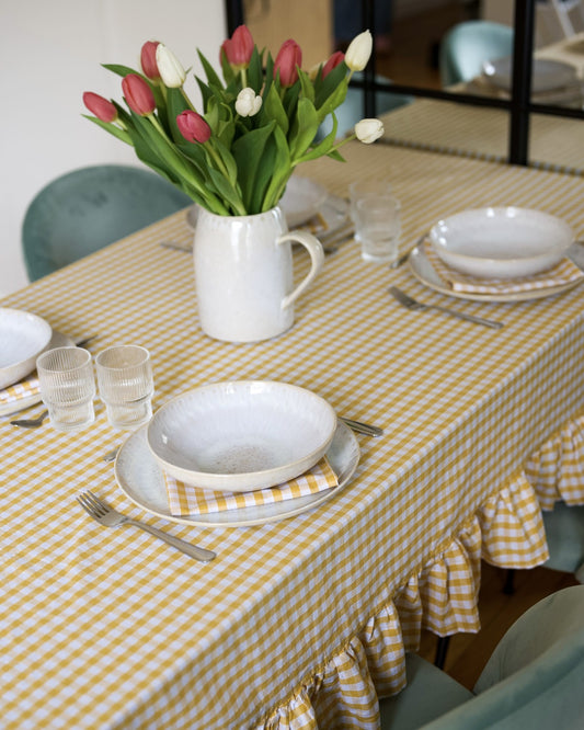 Gros plan sur une nappe à carreaux jaune et blanc avec volants, en coton 100 % fait main à Biarritz, dressée avec une vaisselle artisanale et des serviettes assorties. Au centre, un bouquet de tulipes rouges et blanches dans un pichet en céramique blanche.