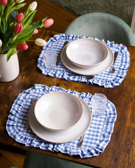 Set de table à carreaux vichy bleu et blanc, en coton 100 %, avec finition matelassée et volant autour. Fabriqué artisanalement à Biarritz, il apporte une touche chic et accueillante à une table soigneusement dressée.
