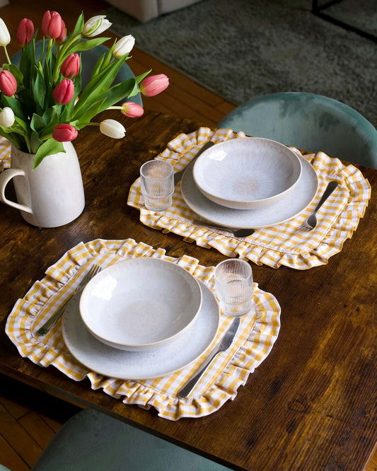 Set de table à carreaux vichy jaune et blanc, en coton 100 % avec finition matelassée et volant sur tout le contour. Fabriqué à la main à Biarritz, il apporte une touche raffinée et chaleureuse à une table joliment dressée.