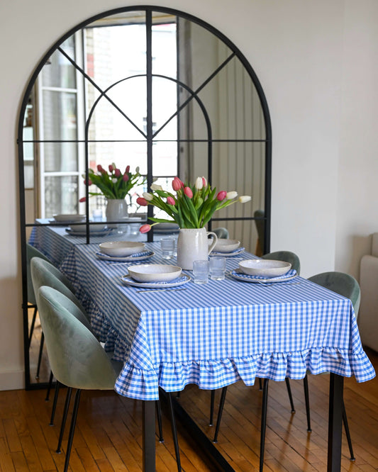 Nappe à carreaux bleu et blanc avec volants, en coton 100 % fabriquée à la main à Biarritz, dressée sur une grande table de salle à manger avec vaisselle en grès et bouquet de tulipes, dans un intérieur lumineux et élégant.