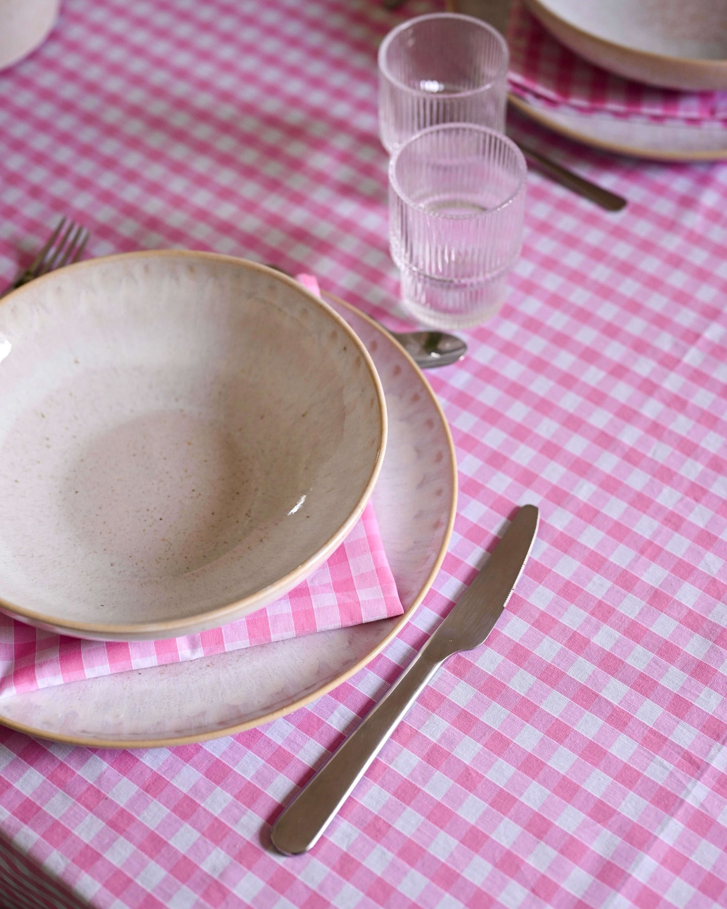 Gros plan sur une nappe à carreaux rose et blanc avec volants, en coton 100 % fait main à Biarritz, dressée avec une vaisselle artisanale et des serviettes assorties. Au centre, un bouquet de tulipes rouges et blanches dans un pichet en céramique blanche.

