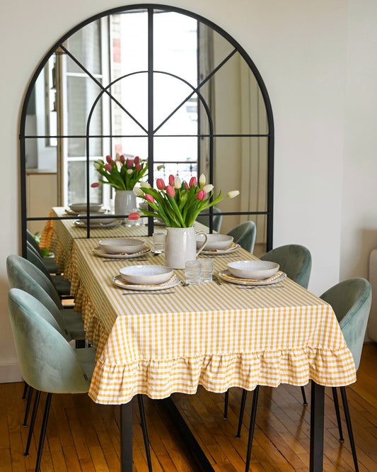 Nappe à carreaux jaune et blanc avec volants, en coton 100 % fabriquée à la main à Biarritz, dressée sur une grande table de salle à manger avec vaisselle en grès et bouquet de tulipes, dans un intérieur lumineux et élégant.
